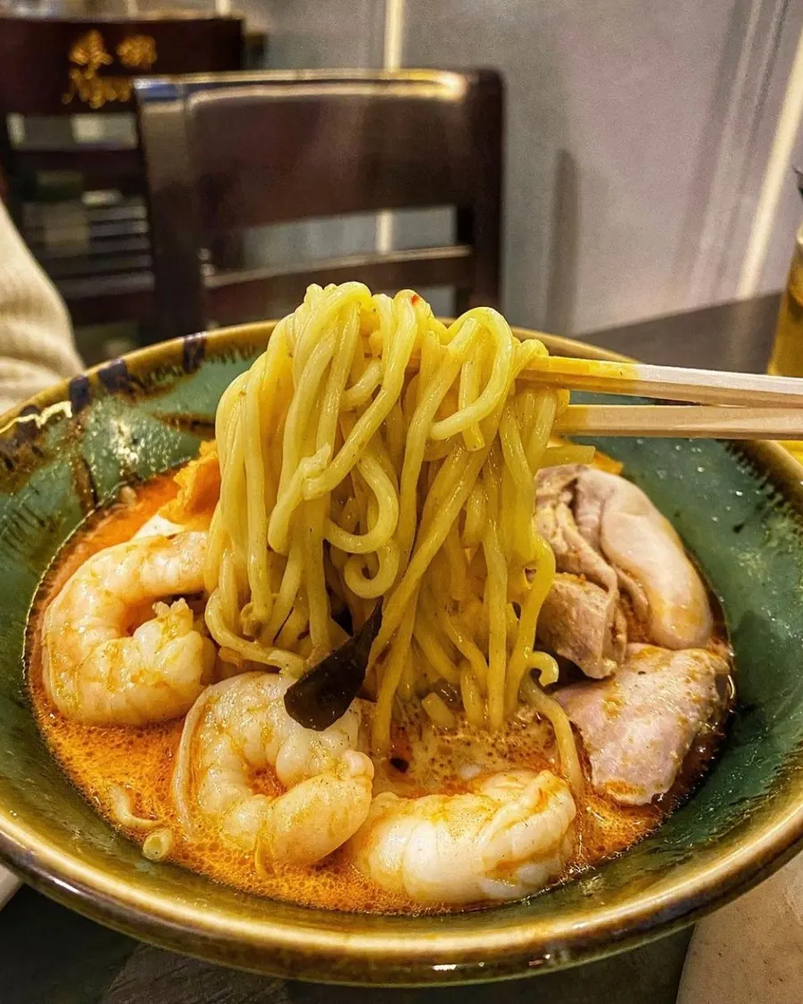 A bowl of laksa curry noodle soup with prawns, noodles lifted with chopsticks